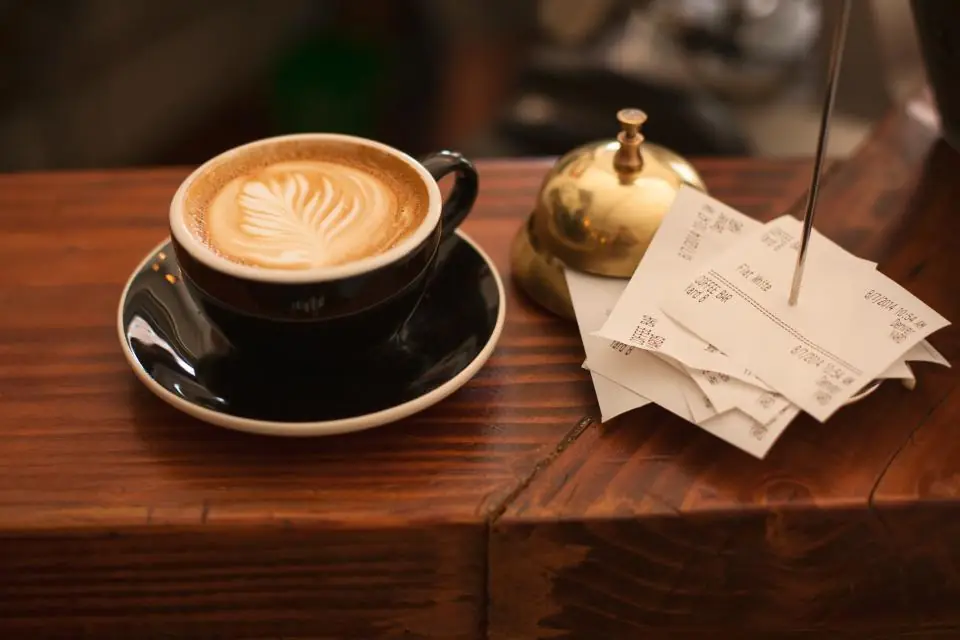 Paper slips on a spike beside a cappuccino and a service bell on a cafe counter