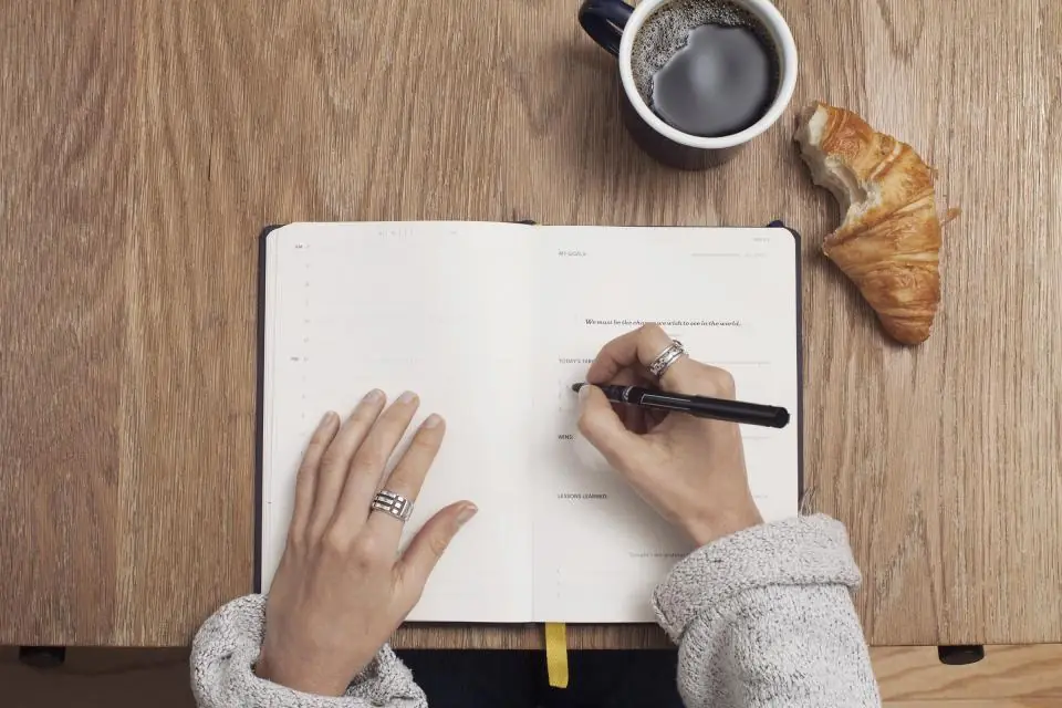 Someone writing a list of questions in a notebook beside a coffee and a croissant